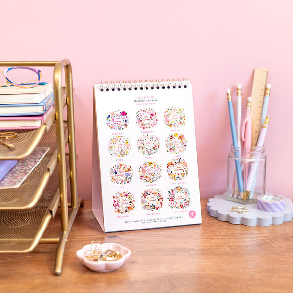 Calendar with floral designs on a desk with stationery items against a pink wall.