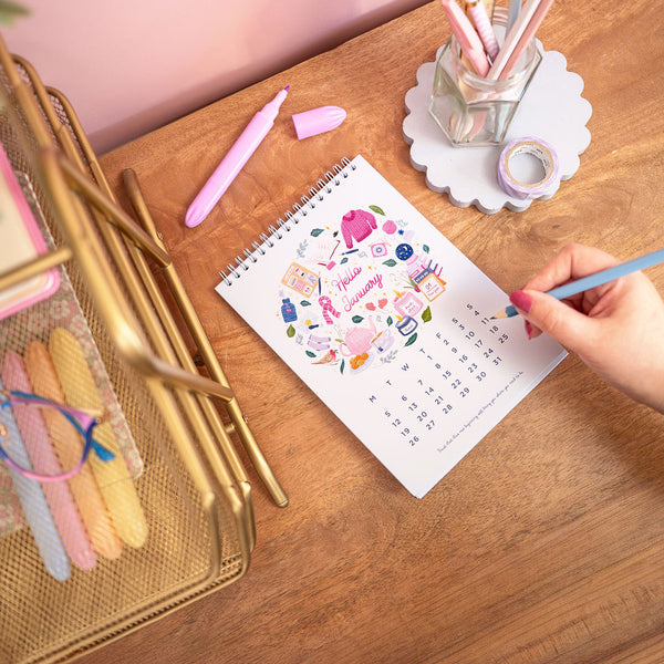 Person using a pen to write on a calendar with decorative elements on a wooden desk.