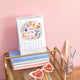 Calendar with 'Hello July' design on a desk with stationery items against a pink background