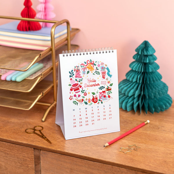 Desk calendar for December on a wooden surface with office supplies and a decorative tree.